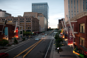 Eine Stadtansicht mit einer Straße voller Autos unten, flankiert von Gebäuden und Wolkenkratzern auf beiden Seiten, unter einem Himmel oben.