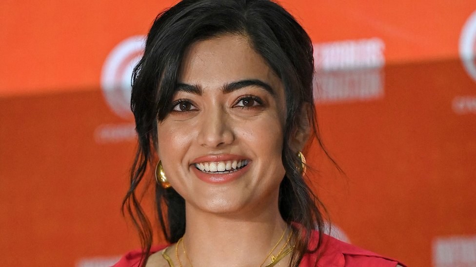 Eine Frau in einem roten Kleid mit einem eleganten Dutt und dezentem Make-up lächelt in die Kamera, trägt schwarze High Heels und hat ein hellgelbes Banner mit der Aufschrift 'Sonam Kapoors Schönste Looks vom Cannes Film Festival' im Hintergrund.
