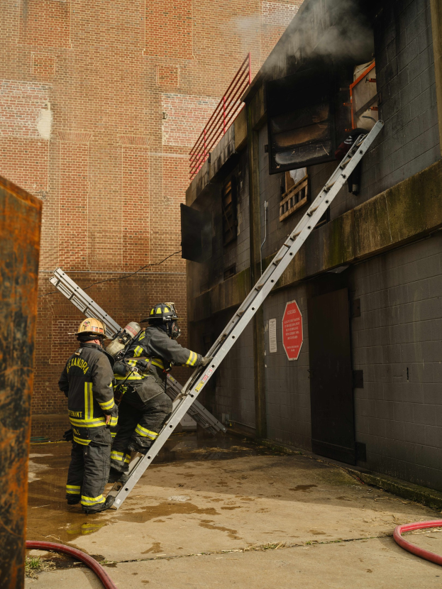 Feuerwehrleute mit Helmen klettern eine Leiter zu einem raucherfüllten Gebäude hinauf, mit einem Metallobjekt links und Rohren unten.
