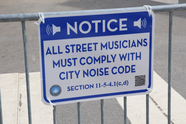 Anschlagtafel an einem Zaun mit einer Tafel, auf der "Hinweis: Alle Straßenmusiker müssen die Stadtlärmvorschrift einhalten" steht, und einer Straße im Hintergrund.