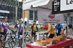 Eine Straßenszene mit Menschen in der Nähe von Fahrrädern, einem Geschäft mit Gegenständen auf einem Tisch, Bäumen, Zelten, an einer Hütte befestigten Schildern, Fahrzeugen und Gras.