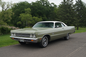 Ein grünes Oldtimer-Auto fährt auf einer Straße mit Bäumen und Wiesen im Hintergrund unter einem klaren Himmel.