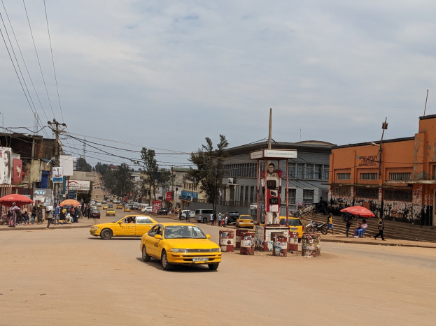 Ein gelbes Taxi fährt eine belebte Stadtstraße mit Gebäuden an den Seiten hinunter, während Fußgänger mit Schirmen auf dem Gehweg unter bewölktem Himmel gehen.