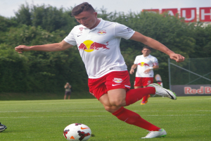 Ein Mann in einem Fußballtrikot kickt einen Ball auf einem grünen Feld mit Bäumen und einem klaren blauen Himmel im Hintergrund, mit einem Schild mit der Aufschrift 'RB Leipzig' im Bild.