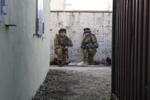 Eine Szene mit einer Wand und einem Fenster auf der linken Seite, einer Eisenwand auf der rechten Seite, zwei Soldaten und Erde in der Mitte sowie Gebäuden, einem Strommast und einer weiteren Wand im Hintergrund.