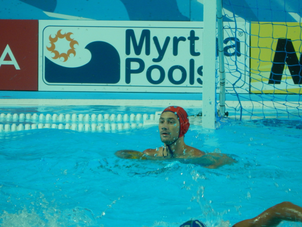 Wasserballspieler in Bewegung in einem Pool, mit einer Mütze tragend, mit einem Netz auf der rechten Seite und einer Werbetafel im Hintergrund.
