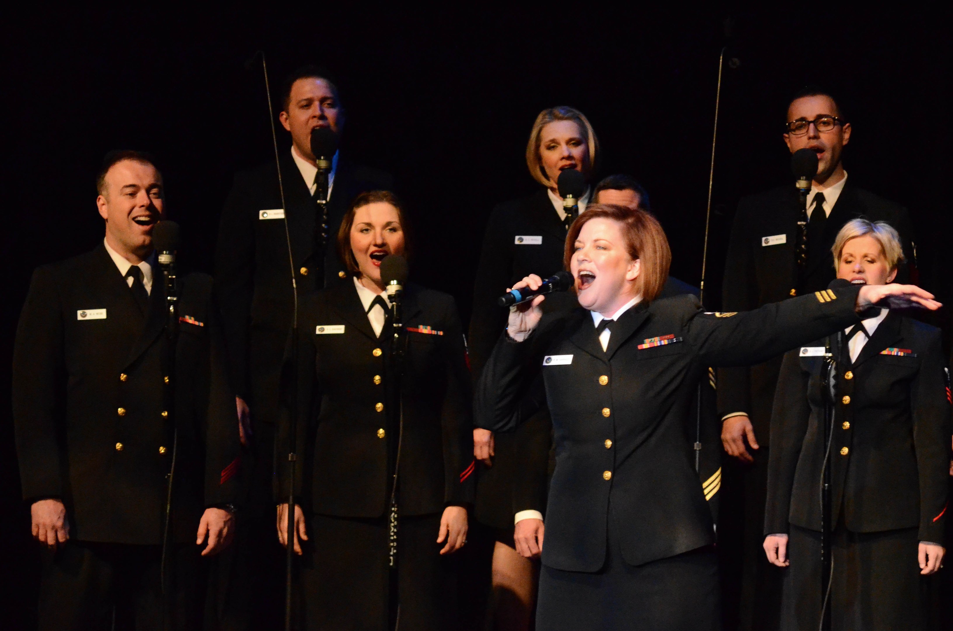 A group of people in military uniforms singing on a stage with microphones and a dark background.