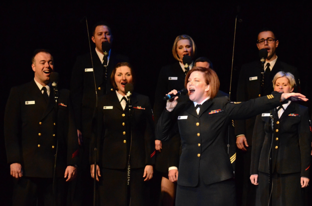 A group of people in military uniforms singing on a stage with microphones and a dark background.