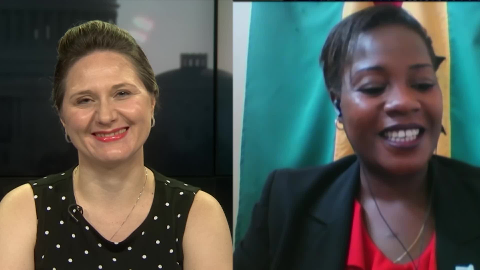 Zwei Frauen, die während eines Videoanrufs lächeln, eine in einem schwarzen und weißen Kleid, die andere in einer unbestimmten Farbe, mit einer Flagge an der Wand hinter ihnen in einer öffentlichen Einrichtung.