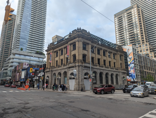 A bustling city street with tall buildings, vehicles, pedestrians, traffic cones, light poles, traffic signals, boards, hoardings, trees, and a cloudy sky, centered on the Toronto Public Library.