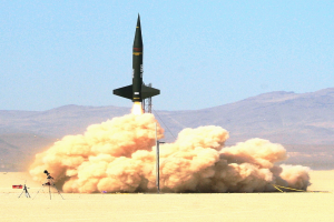 Eine Rakete startet in den Himmel und hinterlässt eine Rauchspur, während Berge und Boden im Hintergrund zu sehen sind.