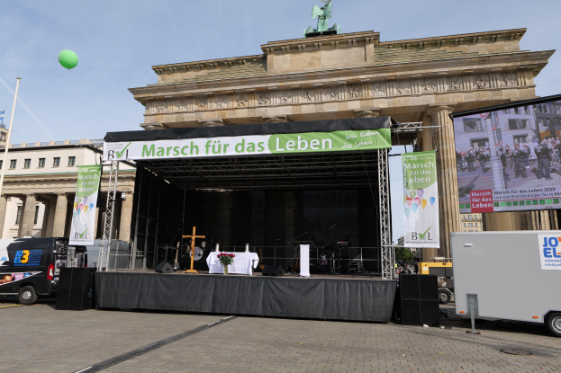 Bühne vor dem Brandenburger Tor mit einem Tisch, Bannern, Lautsprechern, Fahrzeugen, Gebäuden, einer Statue, einer Flagge und Wolken.