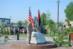 Zwei Militärangehörige halten Fahnen auf beiden Seiten eines Gedenksteins, mit Pflanzen, Autos, anderen Militärangehörigen, Strommasten mit Kabeln und Gebäuden im Hintergrund.