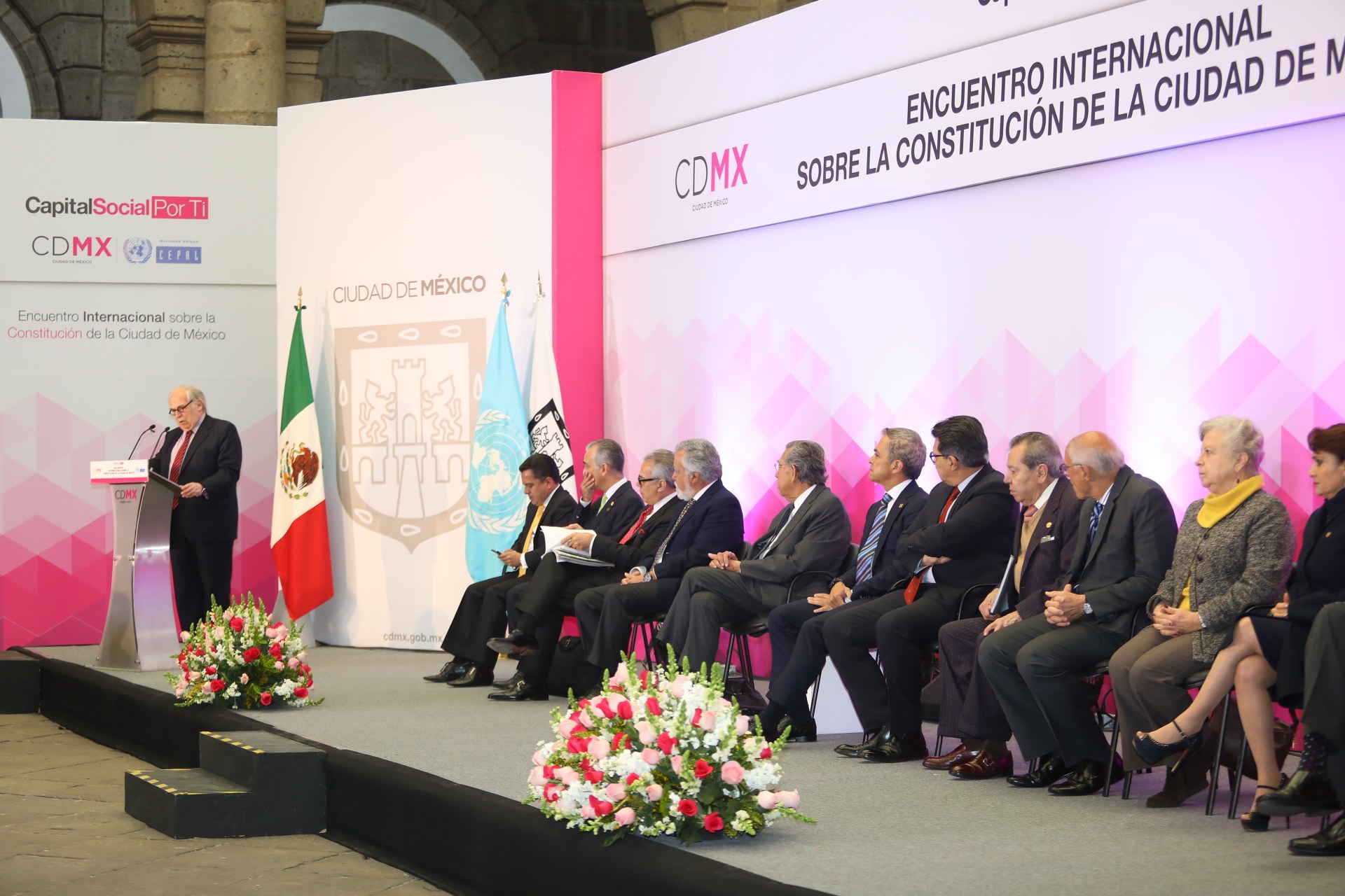Gruppe von Menschen, die auf Stühlen sitzen und einem Podium mit Mikrofon gegenüberstehen, umgeben von Blumenarrangements, Flaggen und Texttafeln in einem Saal mit Säulen und einer Wand, was auf eine Tagung des Internationalen Kongresses von Mexiko hinweist.