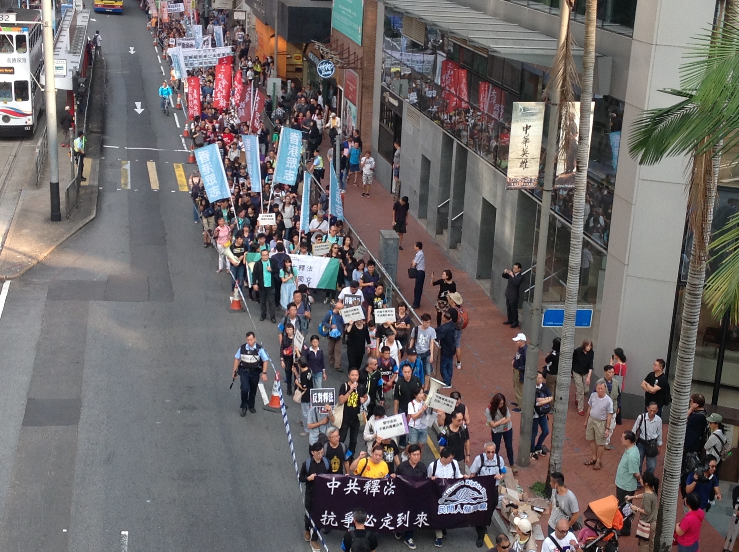 A large group of people marching in a protest down a city street, holding signs and banners, with trees, buildings, and vehicles visible.
