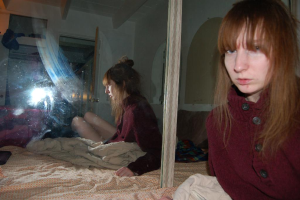 Eine Frau in roter Jacke steht neben einem Spiegel, der ihr Bild reflektiert, mit einem Bett und einer Bettwäsche im Hintergrund.