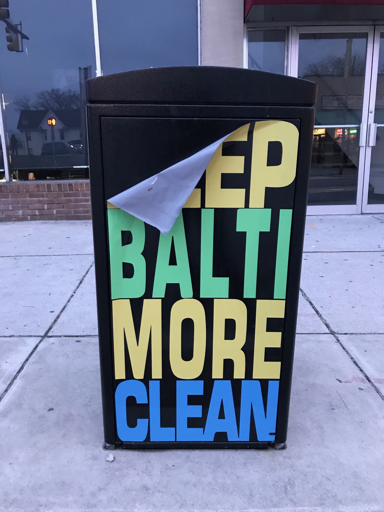 Black trash can with "Keep Baltimore More Clean" text on a sidewalk in front of a building with glass doors.