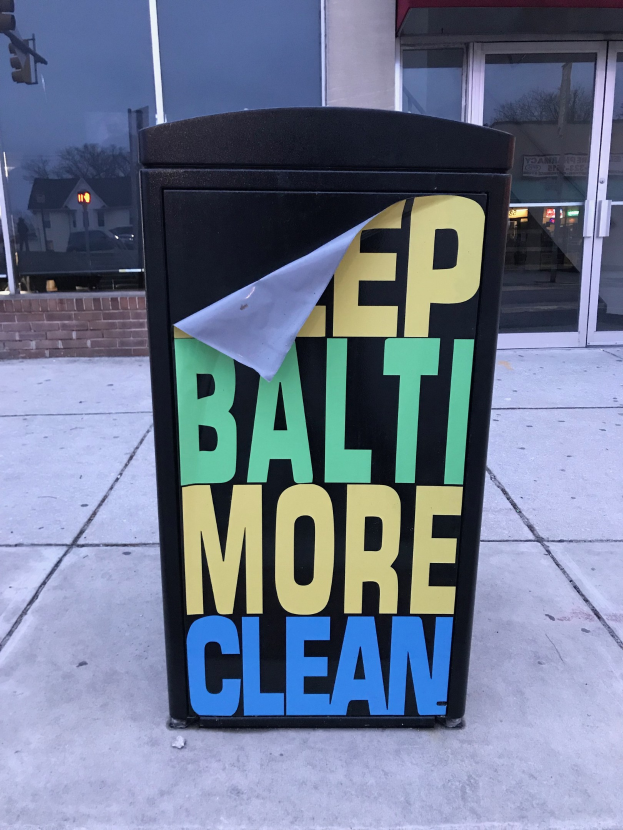 Black trash can with "Keep Baltimore More Clean" text on a sidewalk in front of a building with glass doors.