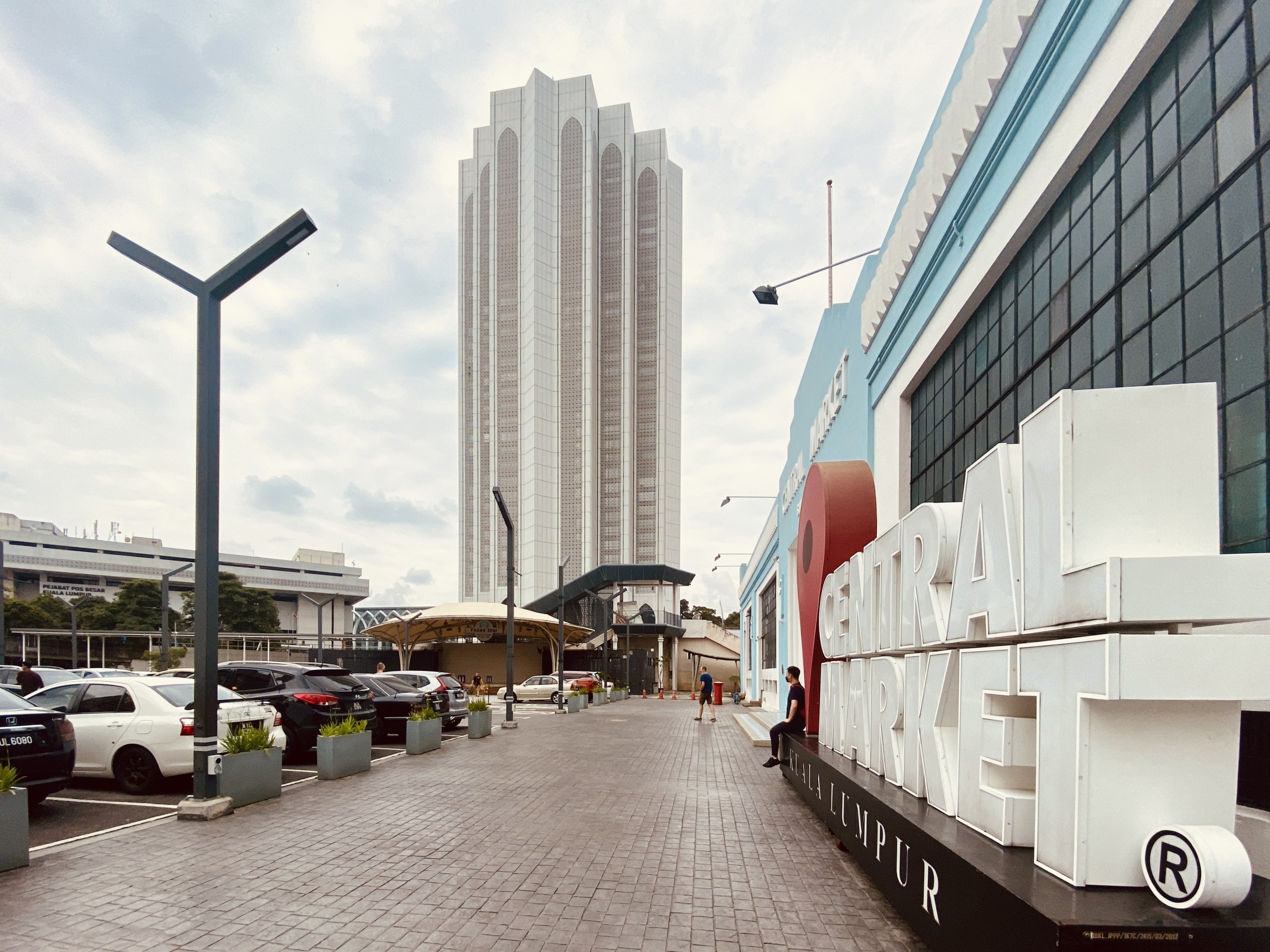 A busy central market in Kuala Lumpur, Malaysia, featuring tall buildings, vehicles, pedestrians, street furniture, signage, trees, and a cloudy sky.