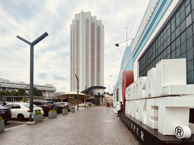 A busy central market in Kuala Lumpur, Malaysia, featuring tall buildings, vehicles, pedestrians, street furniture, signage, trees, and a cloudy sky.