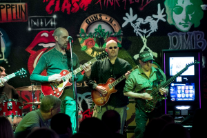 Drei Männer auf einer Bühne mit Gitarren, vor einem Publikum und einer bemalten Wand.