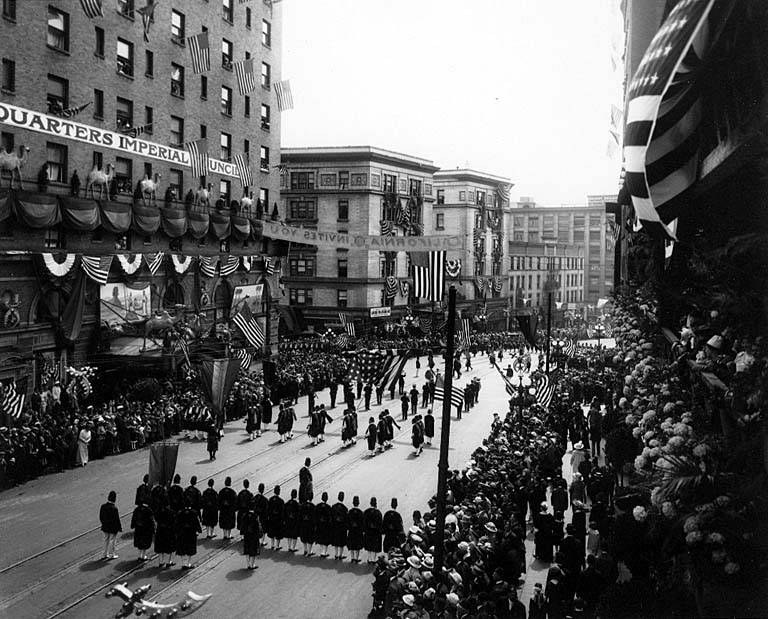 A large crowd of people marches down a New York City street during a Fourth of July parade, many wearing caps and holding flags, with others lining the sides; flags on poles and trees line the street, and buildings with windows and writing in the background are visible under a clear sky.