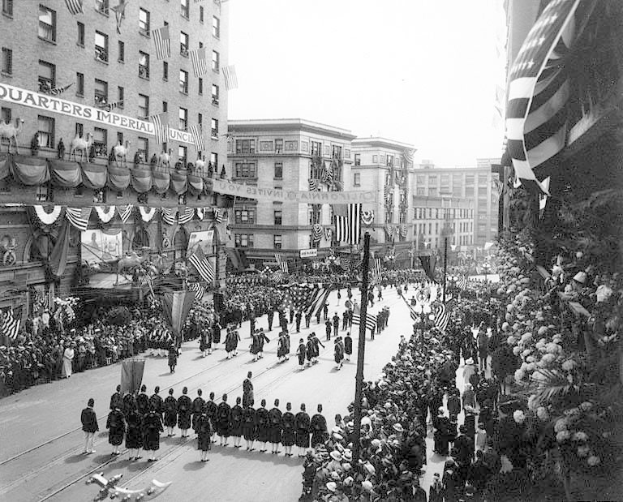 A large crowd of people marches down a New York City street during a Fourth of July parade, many wearing caps and holding flags, with others lining the sides; flags on poles and trees line the street, and buildings with windows and writing in the background are visible under a clear sky.
