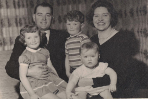 Eine Familie mit fünf Personen, darunter ein Mann in Anzug und Krawatte links, eine Frau rechts und drei Kinder, alle strahlend.