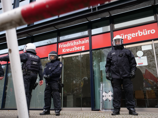 Eine Gruppe von Polizisten in Helmen und Handschuhen steht vor einem Glaswandgebäude, mit einem Pfahl links und Tafeln im Hintergrund, die "Friedrichshain Kreuzberg" und Papiere an den Glaswänden angeklebt zeigen.