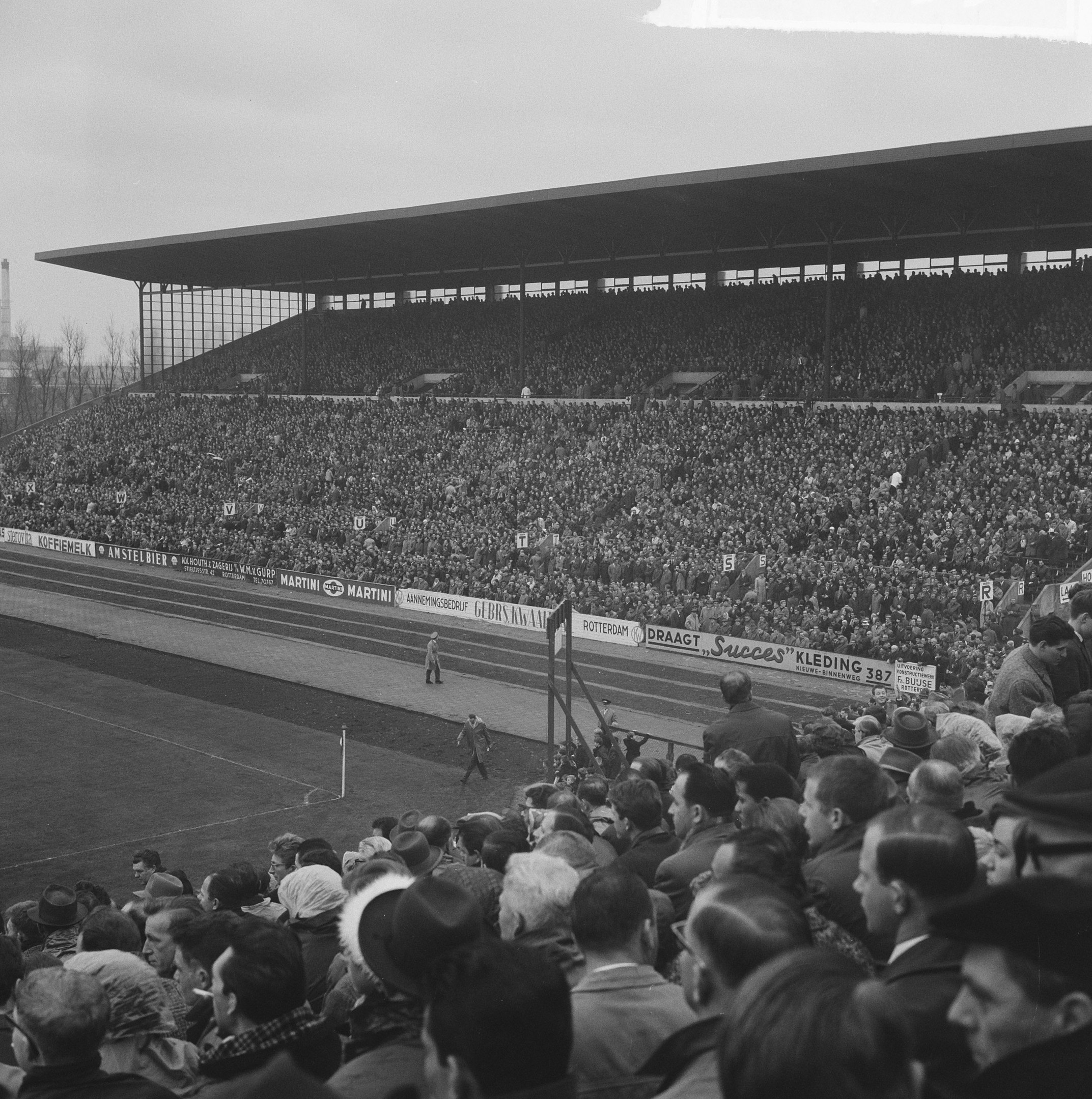 Schwarzes und weißes Foto eines vollen Stadions mit Zuschauern, die ein Fußballspiel verfolgen, mit Bannern, Pfählen, einem Schuppen, Bäumen, einem Turm und einer bewölkten Himmel