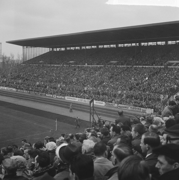 Schwarzes und weißes Foto eines vollen Stadions mit Zuschauern, die ein Fußballspiel verfolgen, mit Bannern, Pfählen, einem Schuppen, Bäumen, einem Turm und einer bewölkten Himmel