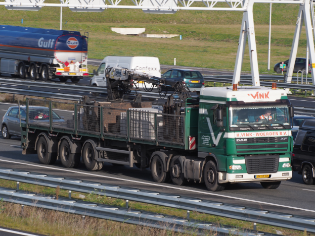 Ein großer Güterzug mit Anhänger fährt auf einer Autobahn neben einer Brücke, mit Gras und Geländern auf beiden Seiten und einem klaren blauen Himmel mit Masten im Hintergrund.