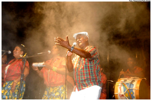 Eine Gruppe von Musikern, die auf der Bühne stehen, mit einem Mann, der in ein Mikrofon singt, und einem anderen Mann, der rechts ein Schlagzeug spielt.