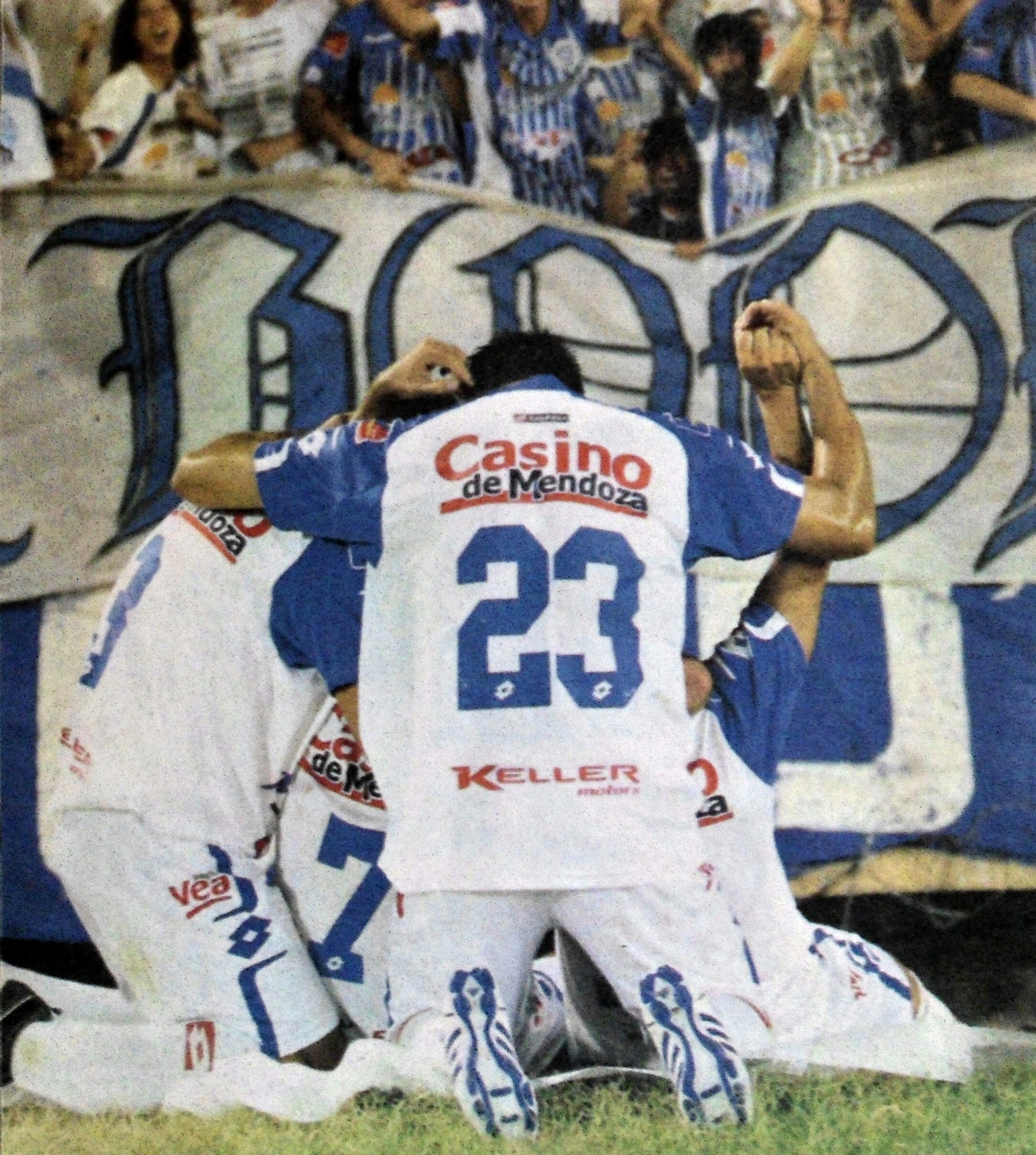 Gruppe von Fußballspielern, die auf dem Platz vor einer jubelnden Menge mit einem Banner im Hintergrund feiern.