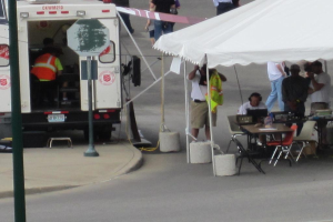 Ein Krankenwagen steht links von einer Straße, während auf der rechten Seite ein Zelt eine Gruppe von Menschen schützt, mit einem Tisch und einem Stuhl in der Nähe.