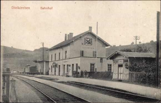 Schwarzes und weißes Foto eines Bahnhofs in Dielkirchen, Deutschland, mit einem Zug auf den Schienen, umgeben von Häusern, Pfählen, Bäumen, Pflanzen und Menschen, Hügel im Hintergrund und klarem Himmel.