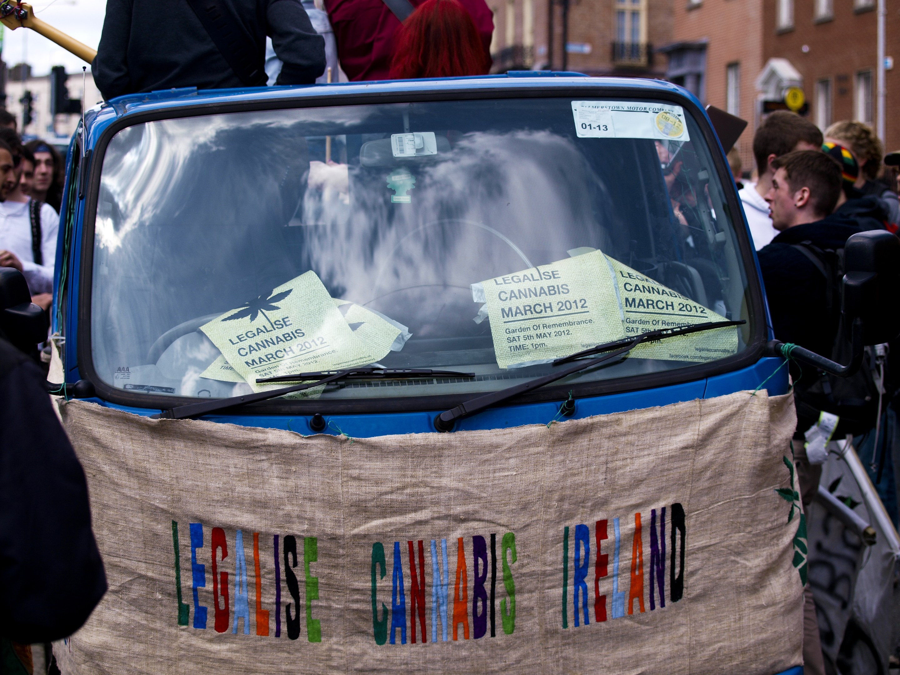 Eine Gruppe von Menschen steht um ein Auto herum, auf dem ein "Legalise Cannabis Ireland"-Schild steht, mit Papieren im Fahrzeug, vor einem Gebäude, Laternenpfählen und einem klaren blauen Himmel im Hintergrund.