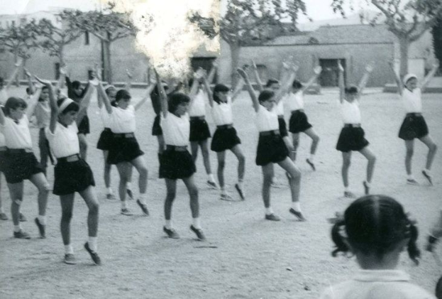 Schwarzes und weißes Bild von jungen Mädchen in Schuluniformen, die auf einer von Gebäuden und Bäumen gesäumten Straße tanzen, mit einer Person in Schuhen unten sichtbar.