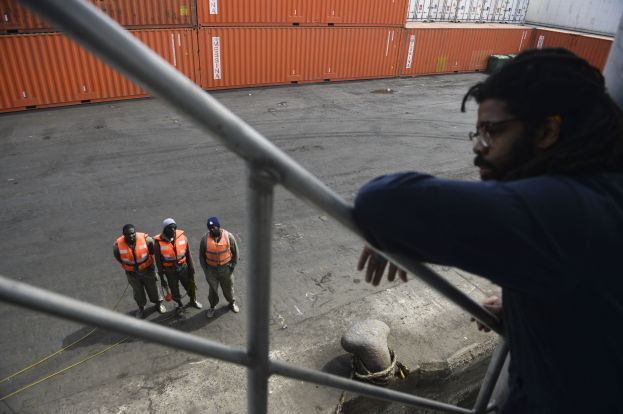 A man in casual clothing stands on a metal railing near a group of people wearing orange jackets and caps, with shipping containers and a wall in the background at the Port of Los Angeles.