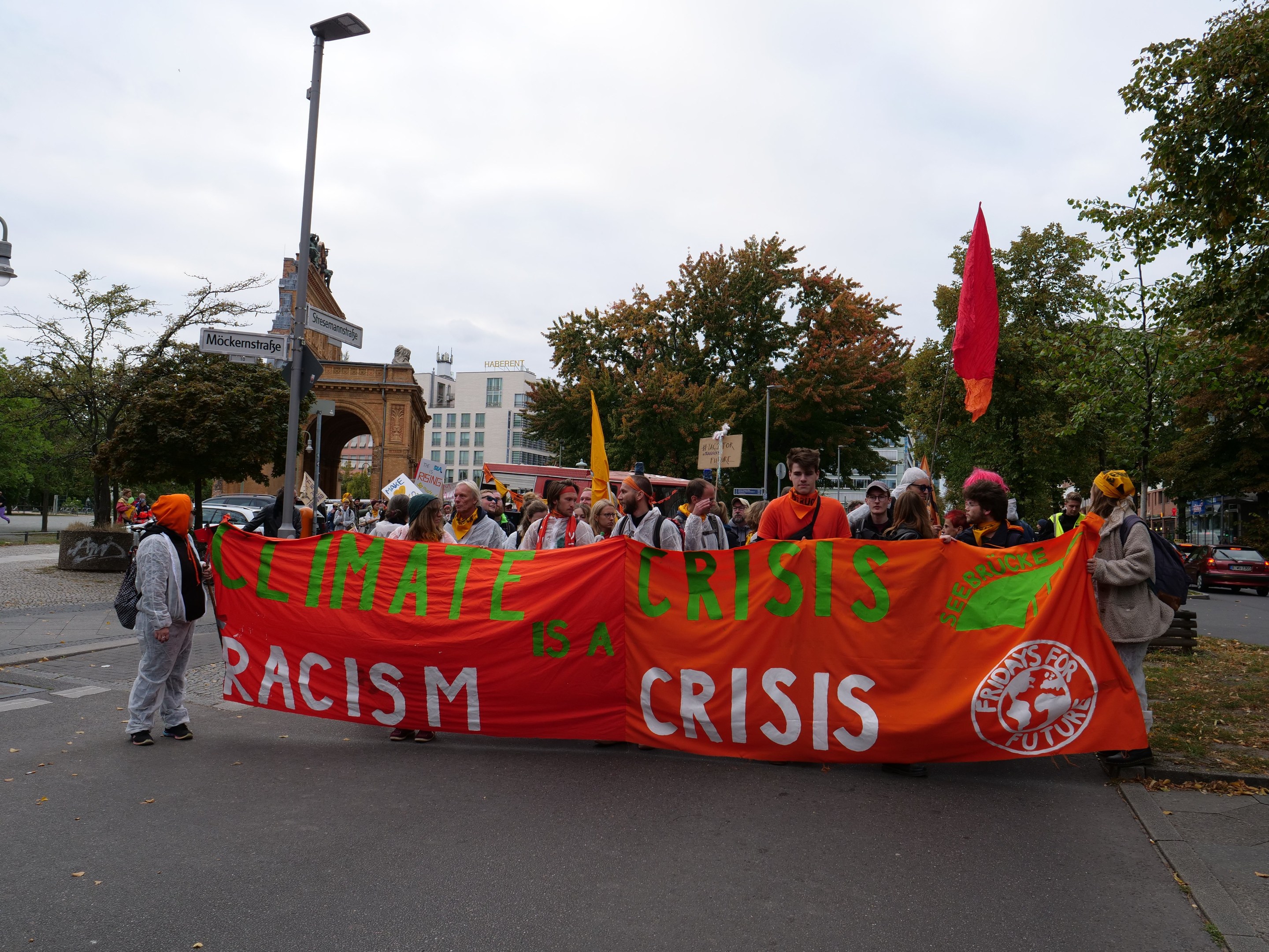 Eine Gruppe von Menschen marschiert eine baumbestandene Straße entlang und hält ein Transparent mit der Aufschrift "Klimakrise ist eine Krise", mit Fahrzeugen und Gebäuden im Hintergrund unter einem klaren blauen Himmel.