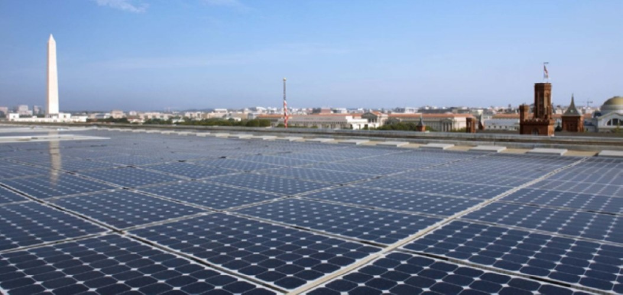 Solarpanels auf dem Dach eines Gebäudes mit dem Washington Monument im Hintergrund, umgeben von Bäumen und anderen Gebäuden unter einem bewölkten Himmel.