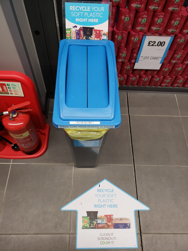 Ein roter Feuerlöscher neben einem blauen Müllcontainer, mit roten Gegenständen auf einer Ablage links und einer weißen Tafel mit der Aufschrift "Recycle your soft plastic right here" rechts.