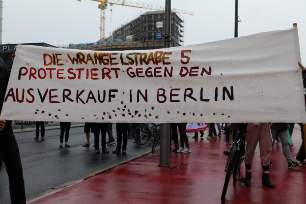Group of people holding a banner reading "Die Wrangellstraße 5 Protestiert Gegen den Ausverkauf in Berlin" on a road with bicycles, poles, buildings, and a crane in the background.