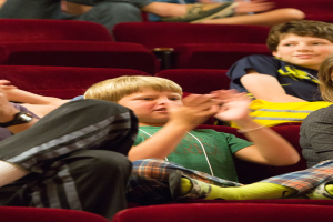 Eine Gruppe von Menschen in einem Kino, die klatschen, mit einem Jungen in der Mitte, umgeben von sitzenden Personen.