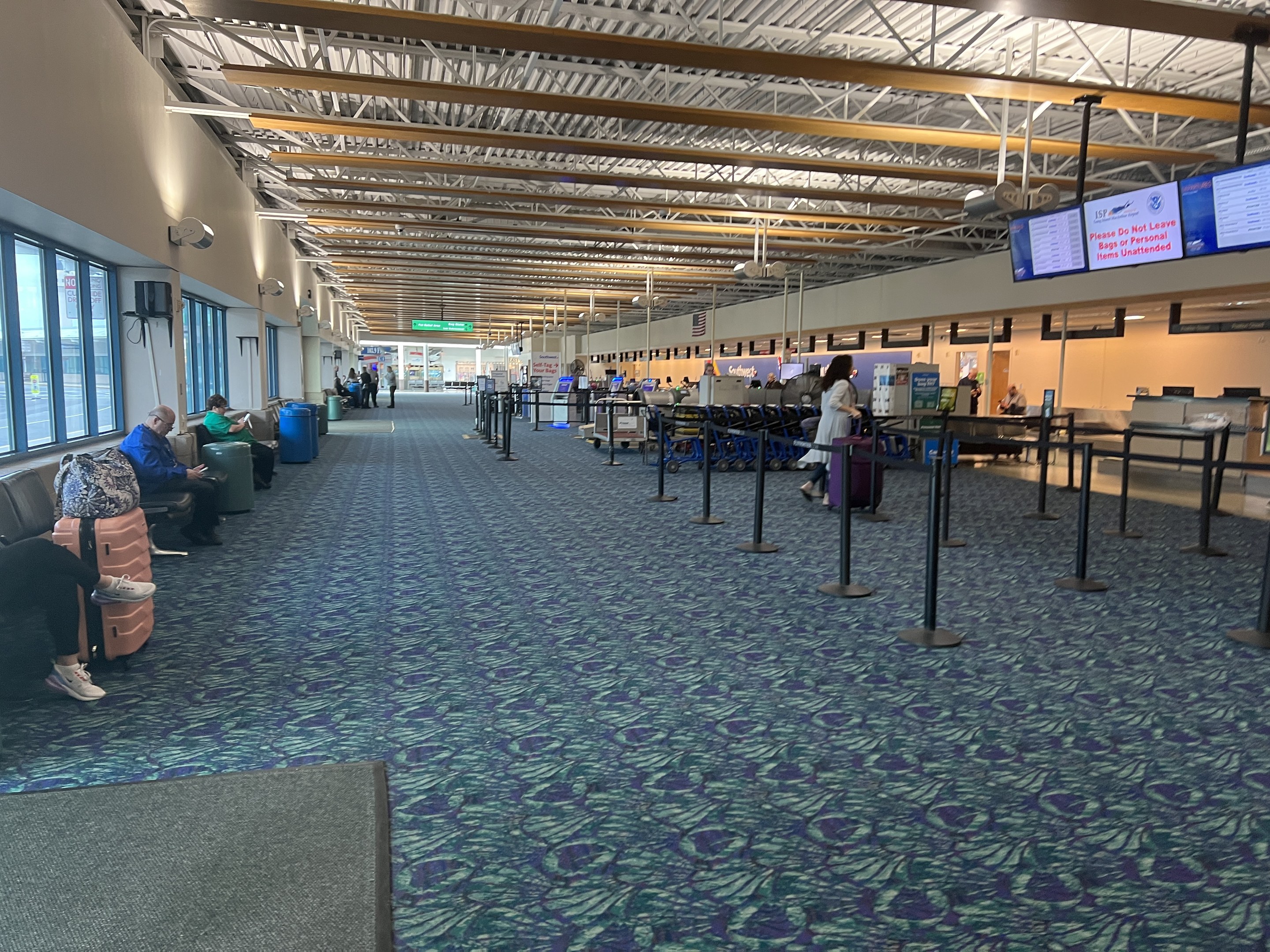 Lange Schlange von Menschen in einem Flughafen-Terminal mit Sitzplätzen, stehenden Passagieren, Bildschirmen an der Wand, Deckenleuchten und Glasfenstern; Hintergrundanzeigen enthalten USDA-Schilder.