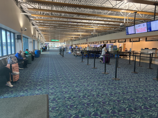 Lange Schlange von Menschen in einem Flughafen-Terminal mit Sitzplätzen, stehenden Passagieren, Bildschirmen an der Wand, Deckenleuchten und Glasfenstern; Hintergrundanzeigen enthalten USDA-Schilder.