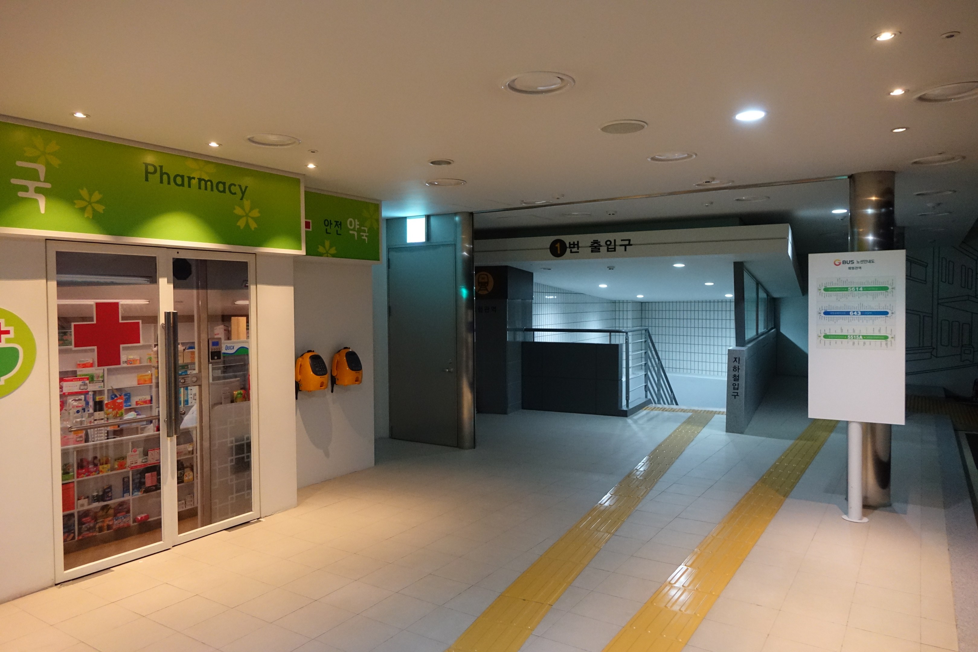 Apothekeneingang mit einem "Apotheke"-Schild auf der linken Seite, einer Tafel mit Text auf der rechten Seite, beleuchteten Lampen oben und undeutlichen Hintergrundobjekten.