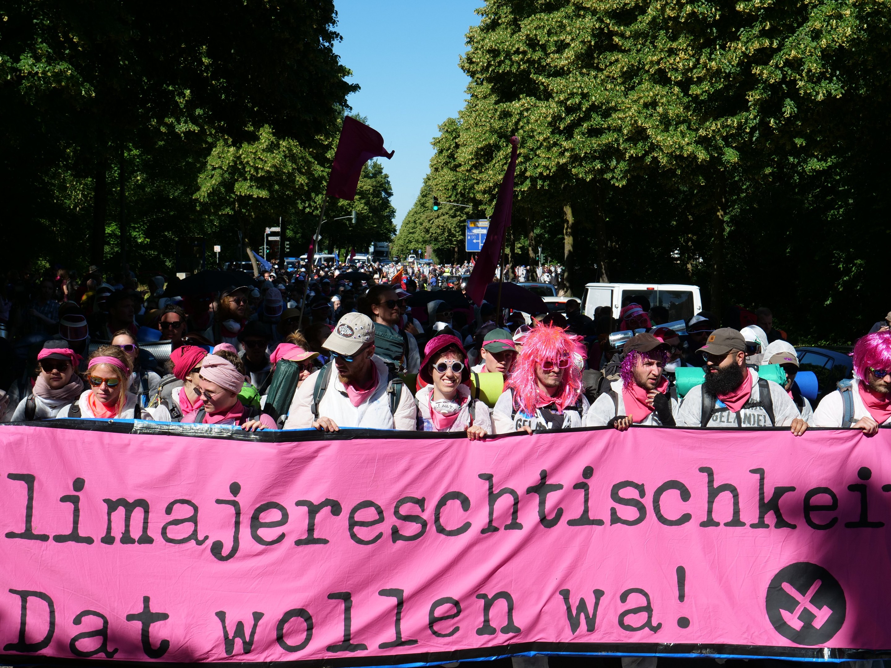 Eine Gruppe von Menschen marschiert die Straße entlang, hält ein pinkes Banner mit der Aufschrift "Women's March in Berlin", einige tragen Mützen und Schutzbrillen, andere halten Fahnen, vor einem Hintergrund aus Bäumen, Fahrzeugen und Himmel.