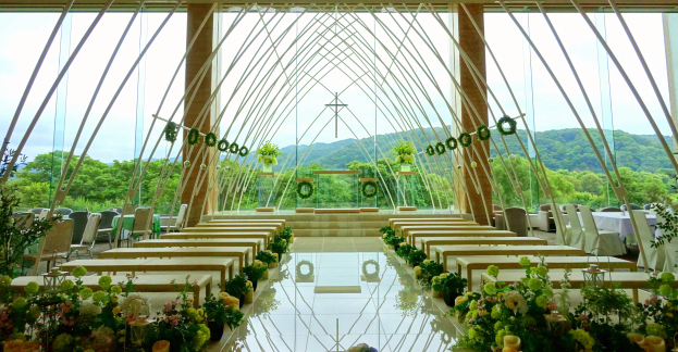Eine Hochzeitszeremonie in einem großen Gebäude mit Tischen und Stühlen in einem Halbkreis angeordnet, geschmückt mit Blumenbouquets, mit Bäumen, Hügeln und einem klaren blauen Himmel im Hintergrund.