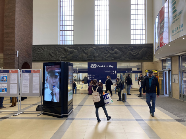 Eine Gruppe von Menschen geht durch einen Bahnhof, einige tragen Taschen, mit Schildern, einem Bildschirm und geschnitzten Wänden im Hintergrund und digitalen Kiosken auf der rechten Seite.